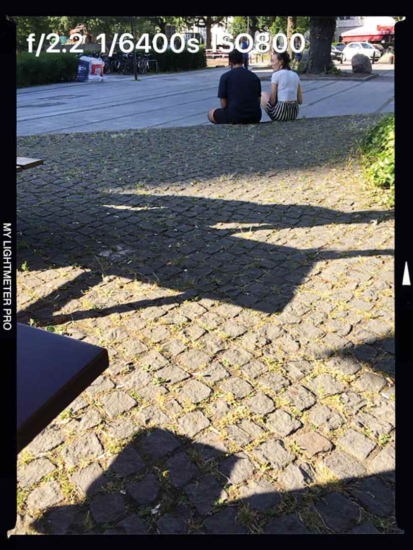 two people sitting on cobblestone street digital production Eine Aufnahme zeigt zwei Personen im Hintergrund auf einem gepflasterten Platz, während sich im Vordergrund Schatten und eine Tischkante befinden. Die Kameraeinstellungen sind f/2.2, 1/6400s Belichtungszeit und ISO 800.