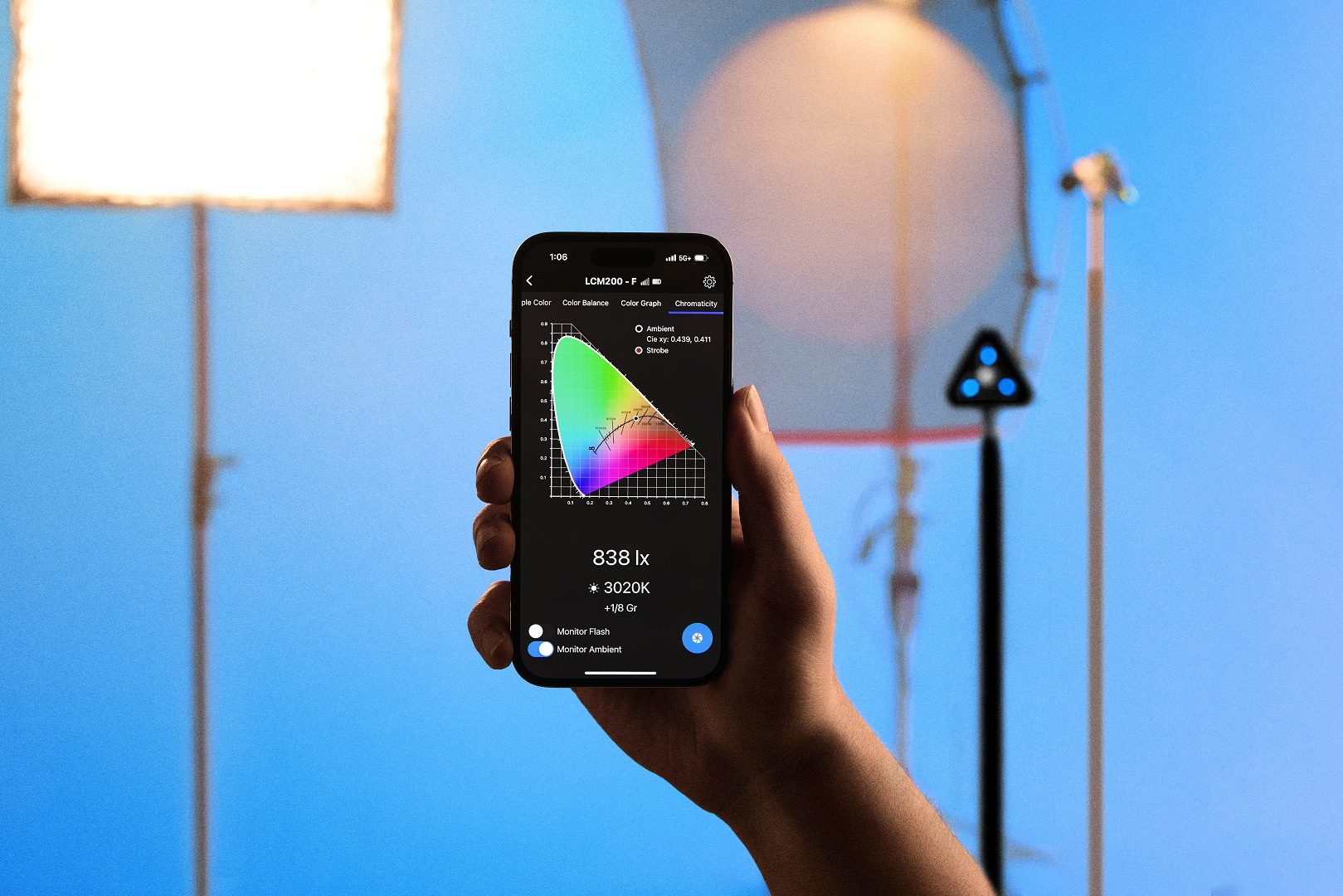 a person holds a smartphone displaying a color spectrum graph in the background are studio lighting equipment and a blue backdrop indicating a professional photography or videography setup digital production A person holds a smartphone displaying a color spectrum graph. In the background are studio lighting equipment and a blue backdrop, indicating a professional photography or videography setup.
