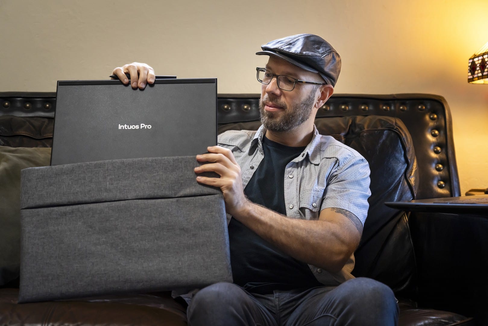a man sitting on a couch holding a drawing tablet in a box labeled intuos pro he is looking at the tablet with an expression of interest the background shows a cozy living room setting with soft lighting digital production A man sitting on a couch holding a drawing tablet in a box labeled 'Intuos Pro.' He is looking at the tablet with an expression of interest. The background shows a cozy living room setting with soft lighting.
