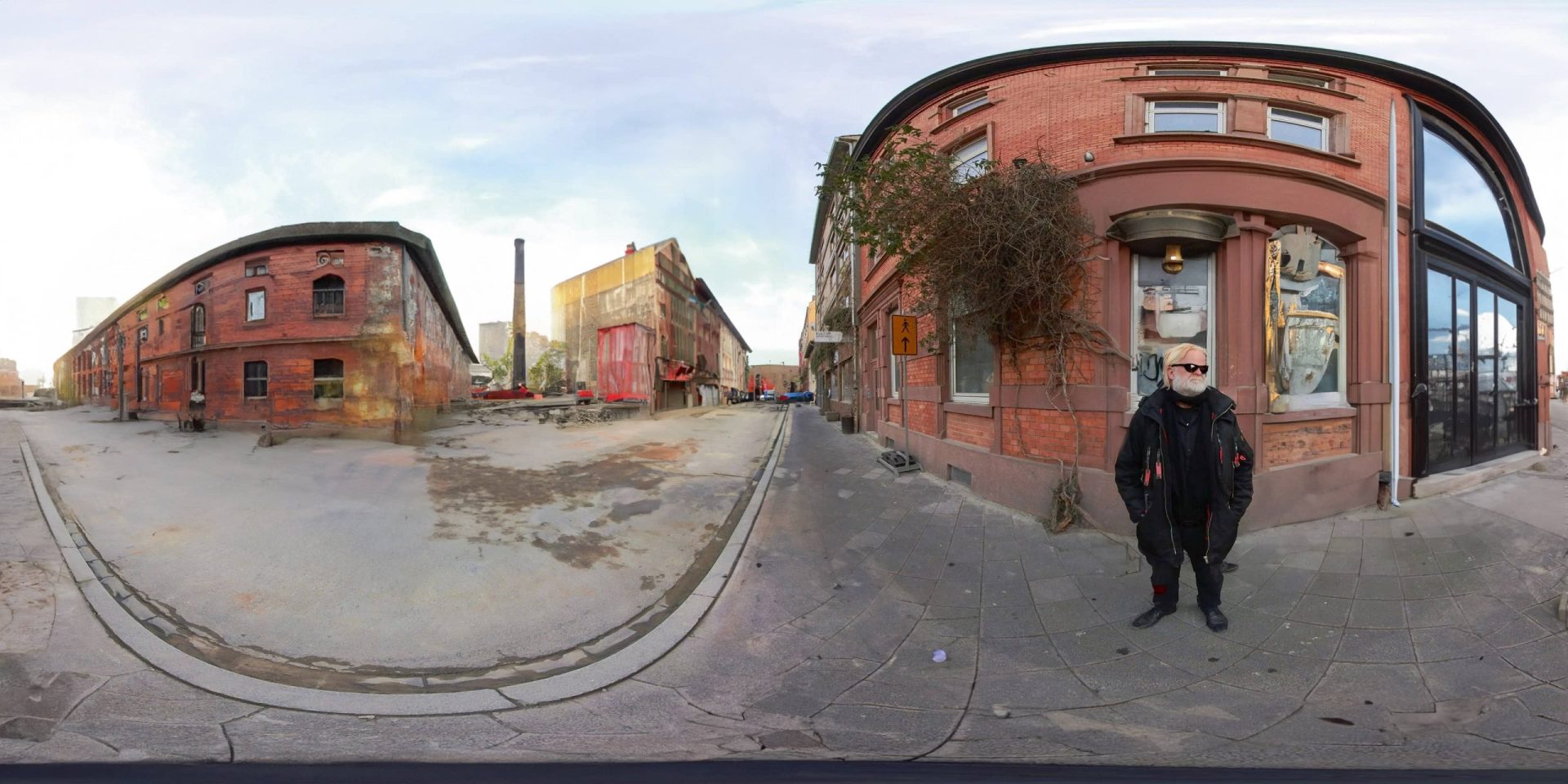 a panoramic view of a street with red brick buildings one showing a storefront with a window a person wearing a black coat and sunglasses is standing on the sidewalk the scene has a cloudy sky and visible industrial elements in the background digital production A panoramic view of a street with red brick buildings, one showing a storefront with a window. A person wearing a black coat and sunglasses is standing on the sidewalk. The scene has a cloudy sky and visible industrial elements in the background.
