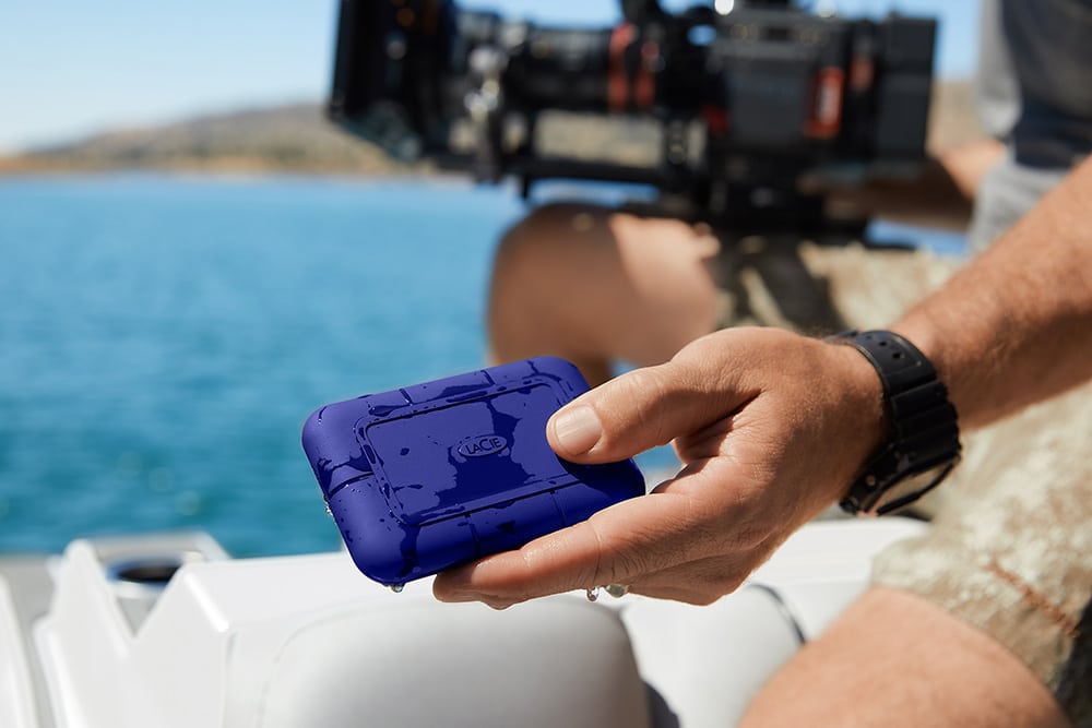 a person holding a blue rugged storage device while sitting on a boat with a camera setup and water in the background digital production A person holding a blue rugged storage device while sitting on a boat, with a camera setup and water in the background.