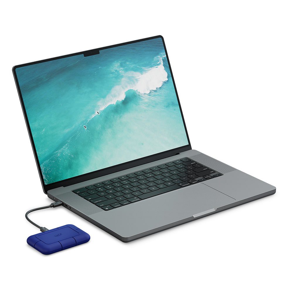 a silver laptop with a display showing an ocean wave connected to a blue external hard drive via a cable the laptop is positioned on a light surface showcasing a modern design digital production A silver laptop with a display showing an ocean wave, connected to a blue external hard drive via a cable. The laptop is positioned on a light surface, showcasing a modern design.
