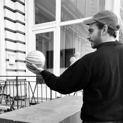 a man in a black sweatshirt and cap holds a striped melon in one hand while standing outside a building with large windows the scene is in black and white featuring a railing and some furniture in the background digital production A man in a black sweatshirt and cap holds a striped melon in one hand while standing outside a building with large windows. The scene is in black and white, featuring a railing and some furniture in the background.