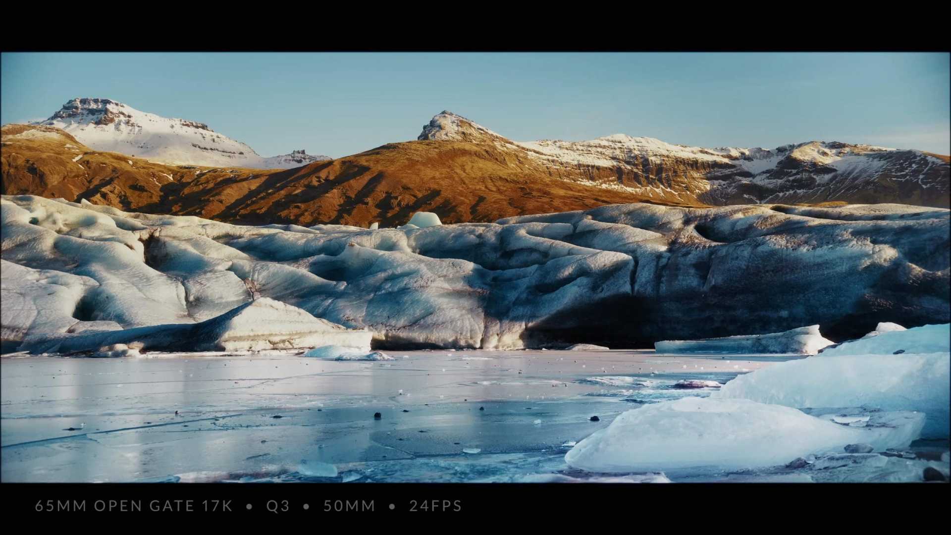 a landscape featuring a glacier and mountain range under a clear sky with patches of snow on the peaks and ice in the foreground text overlay shows technical details about the film format and frame rate digital production A landscape featuring a glacier and mountain range under a clear sky, with patches of snow on the peaks and ice in the foreground. Text overlay shows technical details about the film format and frame rate.