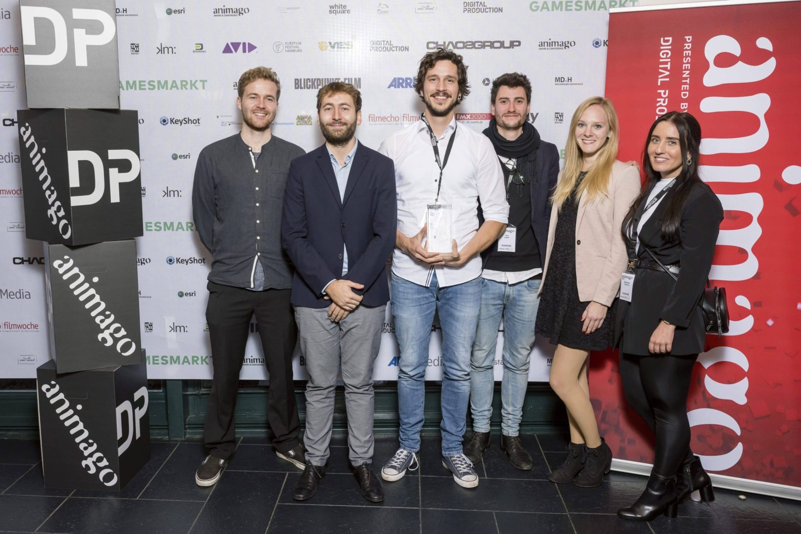 a group of six individuals standing together at an event some holding an award the backdrop features promotional banners for dp and animago everyone is dressed in business casual attire smiling at the camera digital production A group of six individuals standing together at an event, some holding an award. The backdrop features promotional banners for 'DP' and 'animago'. Everyone is dressed in business casual attire, smiling at the camera.