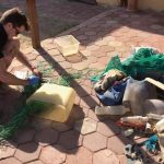 a person crouching on a patio sorting various pieces of plastic waste and netting into piles sunlight casts shadows on the ground highlighting the disarray of discarded materials digital production A person crouching on a patio, sorting various pieces of plastic waste and netting into piles. Sunlight casts shadows on the ground, highlighting the disarray of discarded materials.