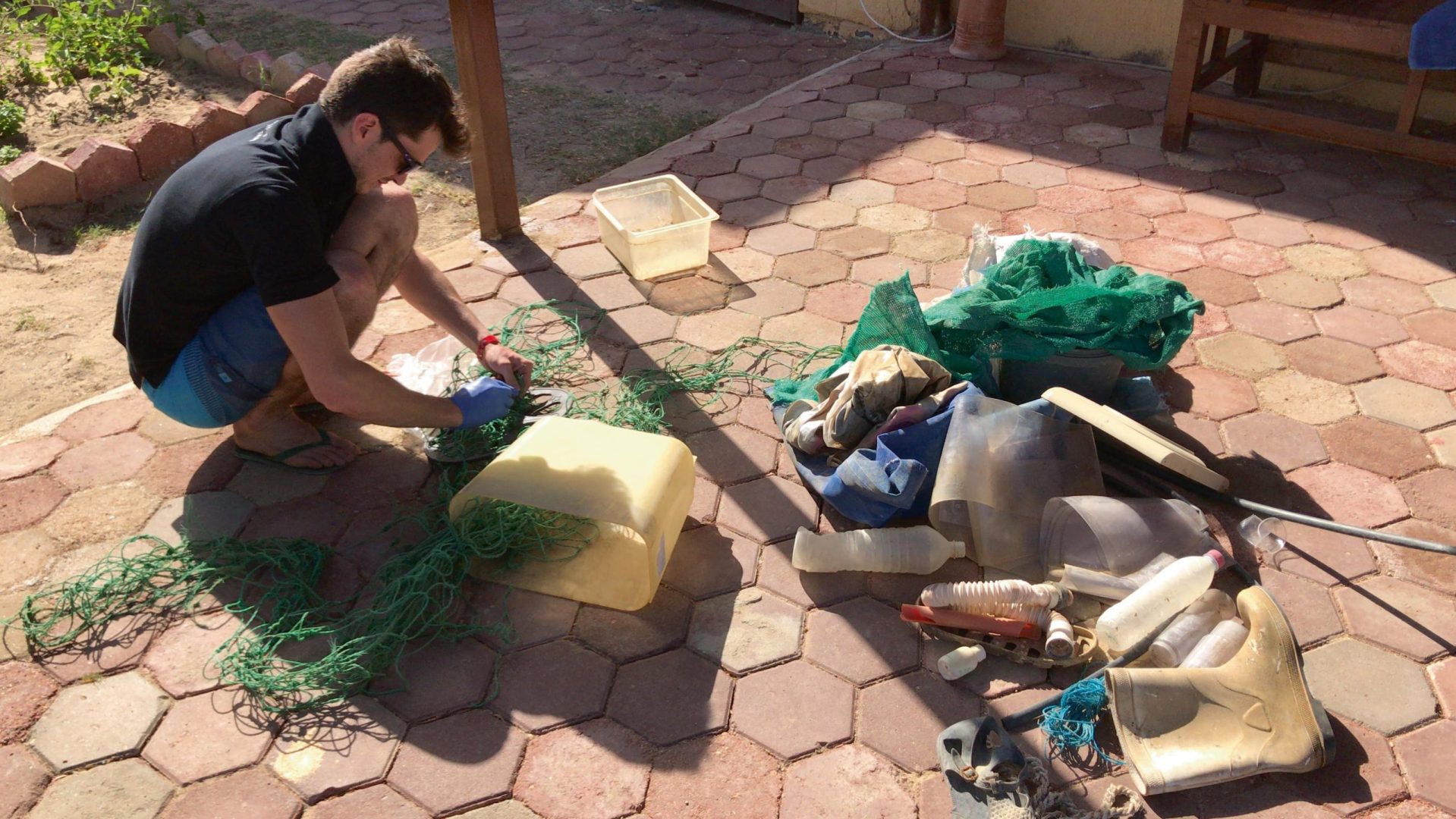 a person crouching on a patio sorting various pieces of plastic waste and netting into piles sunlight casts shadows on the ground highlighting the disarray of discarded materials digital production A person crouching on a patio, sorting various pieces of plastic waste and netting into piles. Sunlight casts shadows on the ground, highlighting the disarray of discarded materials.