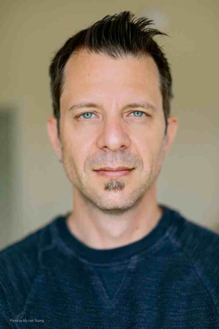 a man with short styled hair and a goatee is looking directly at the camera wearing a dark blue sweater he has light blue eyes and a neutral expression the background is soft and out of focus digital production A man with short, styled hair and a goatee is looking directly at the camera, wearing a dark blue sweater. He has light blue eyes and a neutral expression. The background is soft and out of focus.