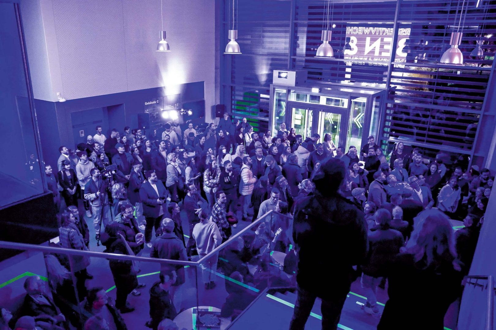 a large crowd of people gathered in a spacious event area with colorful lighting the scene shows attendees socializing with a person standing on a staircase overlooking the crowd digital production A large crowd of people gathered in a spacious event area with colorful lighting. The scene shows attendees socializing, with a person standing on a staircase overlooking the crowd.