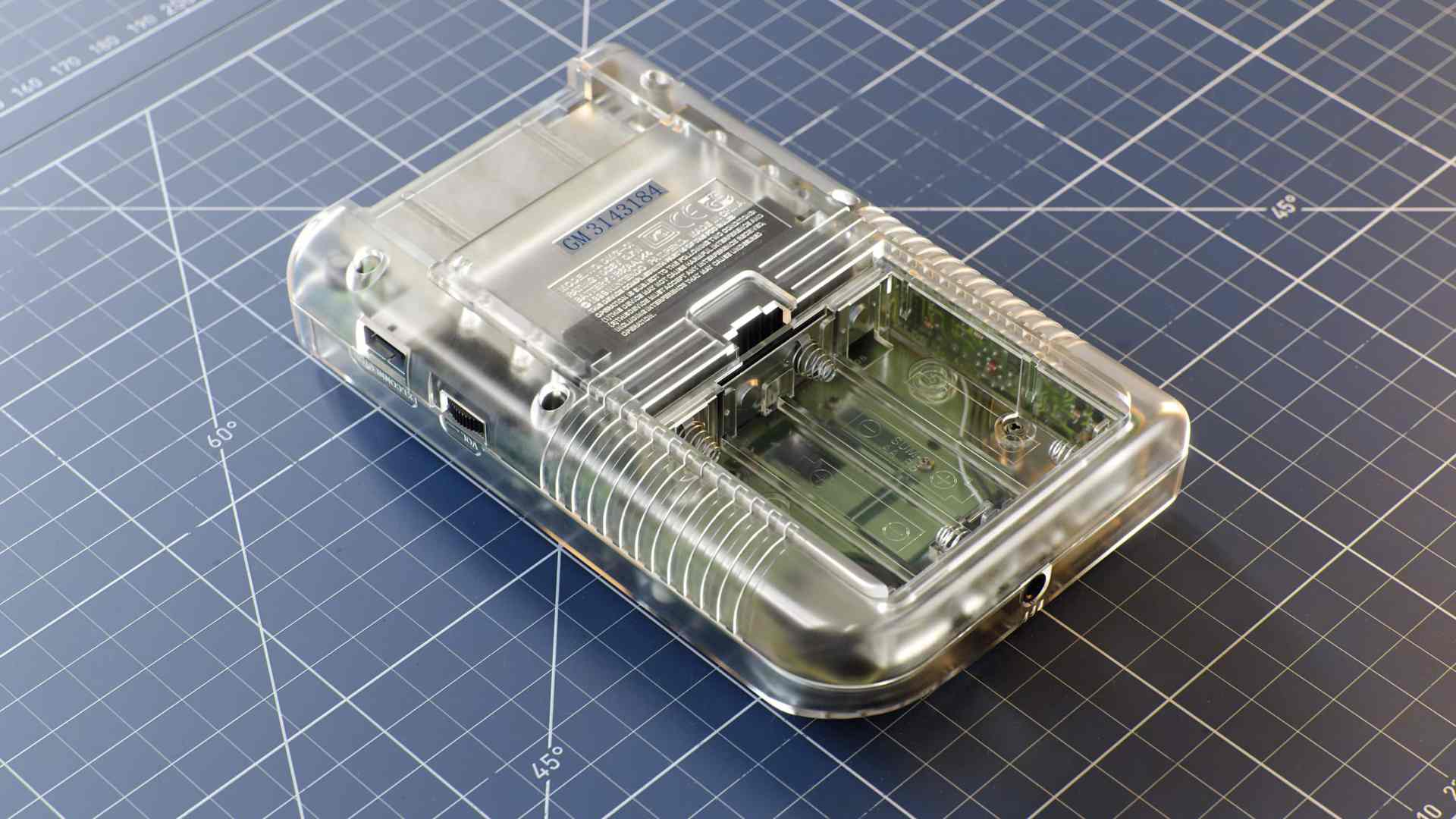 a clear handheld gaming device with visible internal components positioned on a gray grid cutting mat with measurements the device has buttons and ports for connectivity digital production A clear handheld gaming device with visible internal components, positioned on a gray grid cutting mat with measurements. The device has buttons and ports for connectivity.