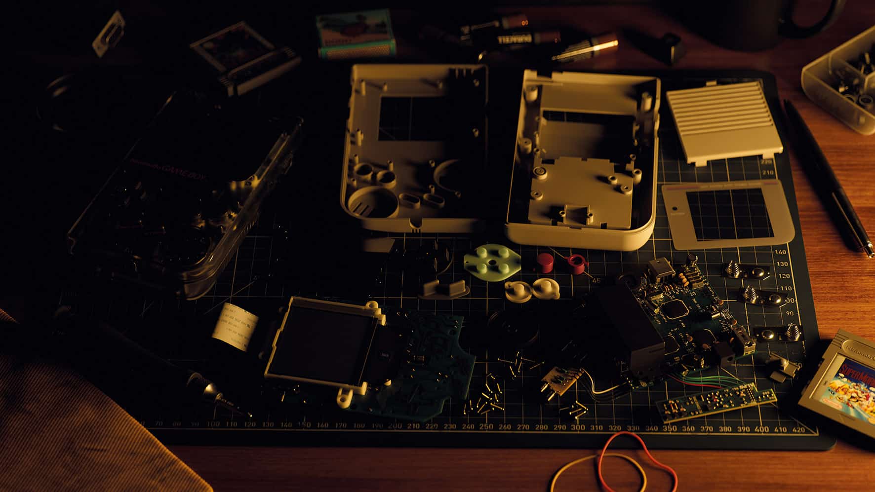 an overhead view of a wooden worktable cluttered with disassembled electronic components including circuit boards batteries screws and tools the lighting is dim highlighting the parts scattered on a cutting mat digital production An overhead view of a wooden worktable cluttered with disassembled electronic components, including circuit boards, batteries, screws, and tools. The lighting is dim, highlighting the parts scattered on a cutting mat.