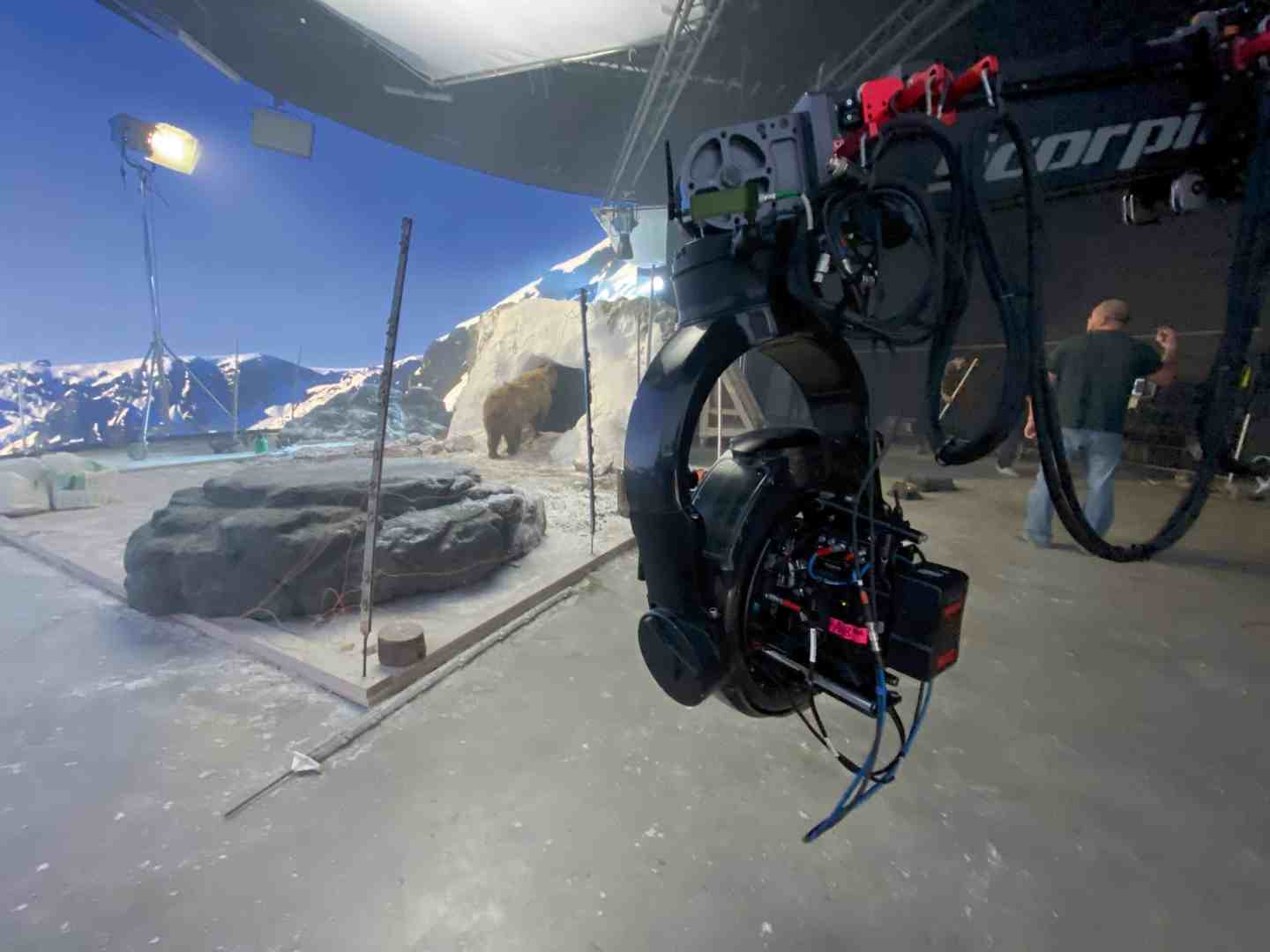 a camera rig mounted on a gimbal positioned in a studio with a backdrop of mountains and a rocky area crew members can be seen working in the background under studio lights digital production A camera rig mounted on a gimbal, positioned in a studio with a backdrop of mountains and a rocky area. Crew members can be seen working in the background under studio lights.
