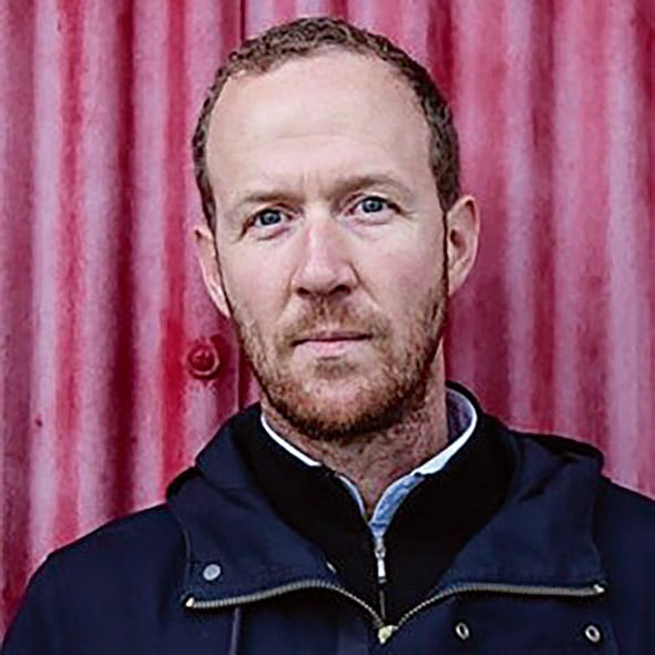 a man with short hair and a beard wearing a dark jacket stands against a textured red wall his expression is neutral and he looks directly at the camera digital production A man with short hair and a beard, wearing a dark jacket, stands against a textured red wall. His expression is neutral and he looks directly at the camera.