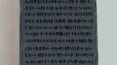 a vertical rectangular stone tablet featuring ancient inscriptions in a series of lines and symbols the tablet is dark gray and rests on a light surface showcasing its engraved markings clearly digital production A vertical, rectangular stone tablet featuring ancient inscriptions in a series of lines and symbols. The tablet is dark gray and rests on a light surface, showcasing its engraved markings clearly.