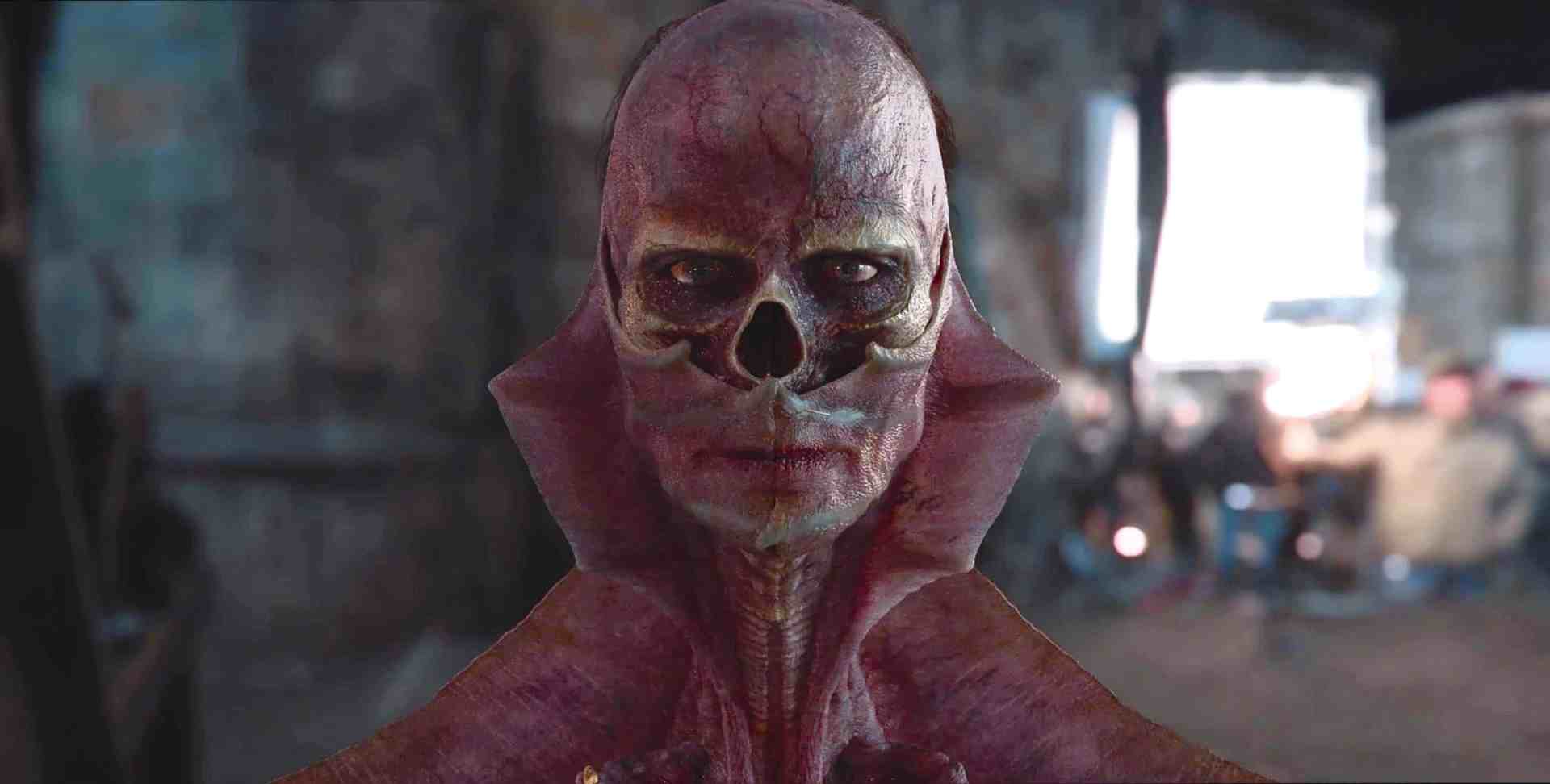 a humanoid figure with a skeletal face and a distinct textured surface is positioned in the foreground of a scene with individuals in the background appearing blurred the setting seems to be dimly lit and rustic digital production A humanoid figure with a skeletal face and a distinct, textured surface is positioned in the foreground of a scene, with individuals in the background appearing blurred. The setting seems to be dimly lit and rustic.