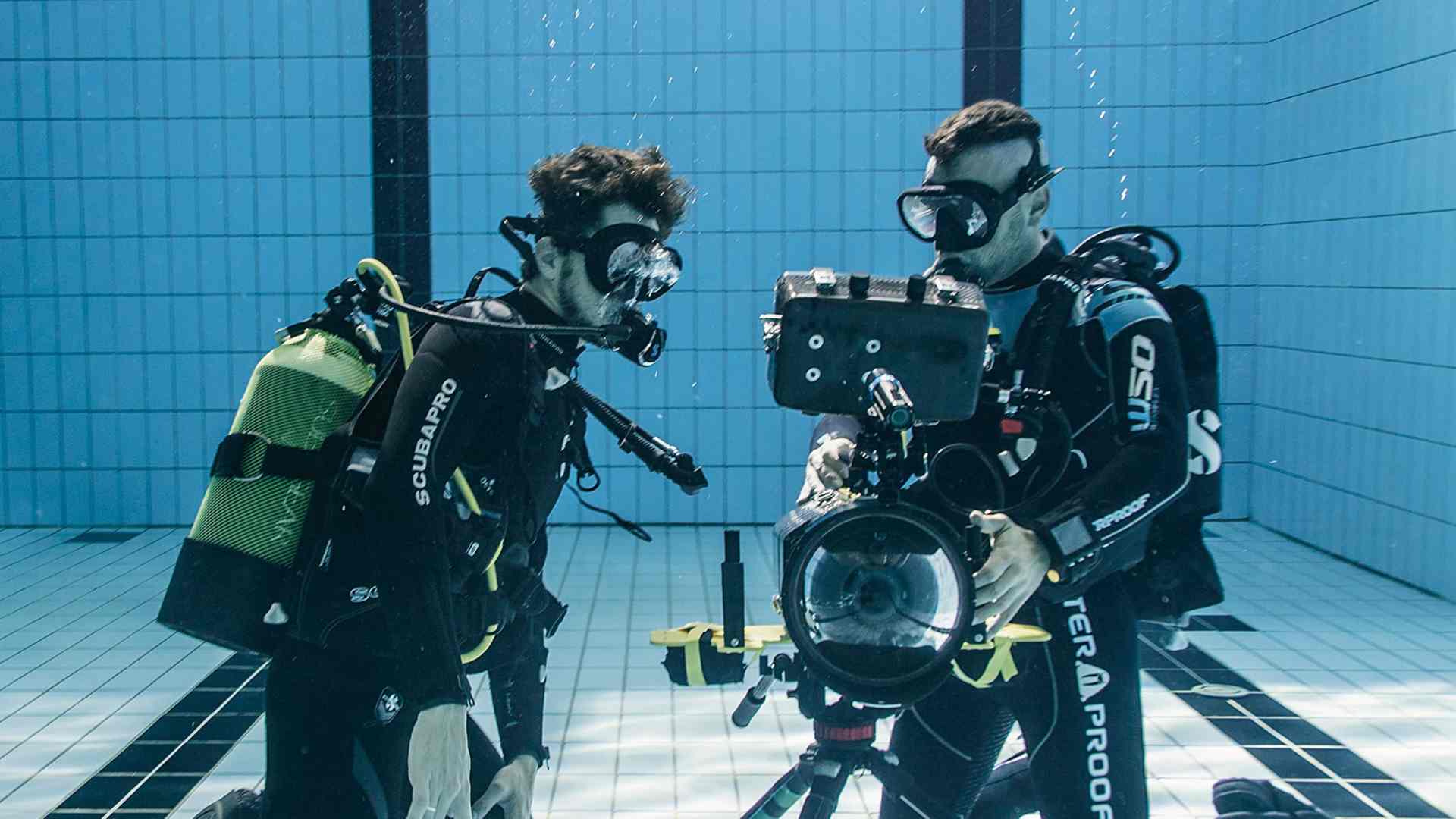 two scuba divers underwater examining underwater filming equipment with a camera visible they are wearing diving suits and accessories surrounded by blue tiled walls of a swimming pool digital production Two scuba divers underwater examining underwater filming equipment, with a camera visible. They are wearing diving suits and accessories, surrounded by blue tiled walls of a swimming pool.