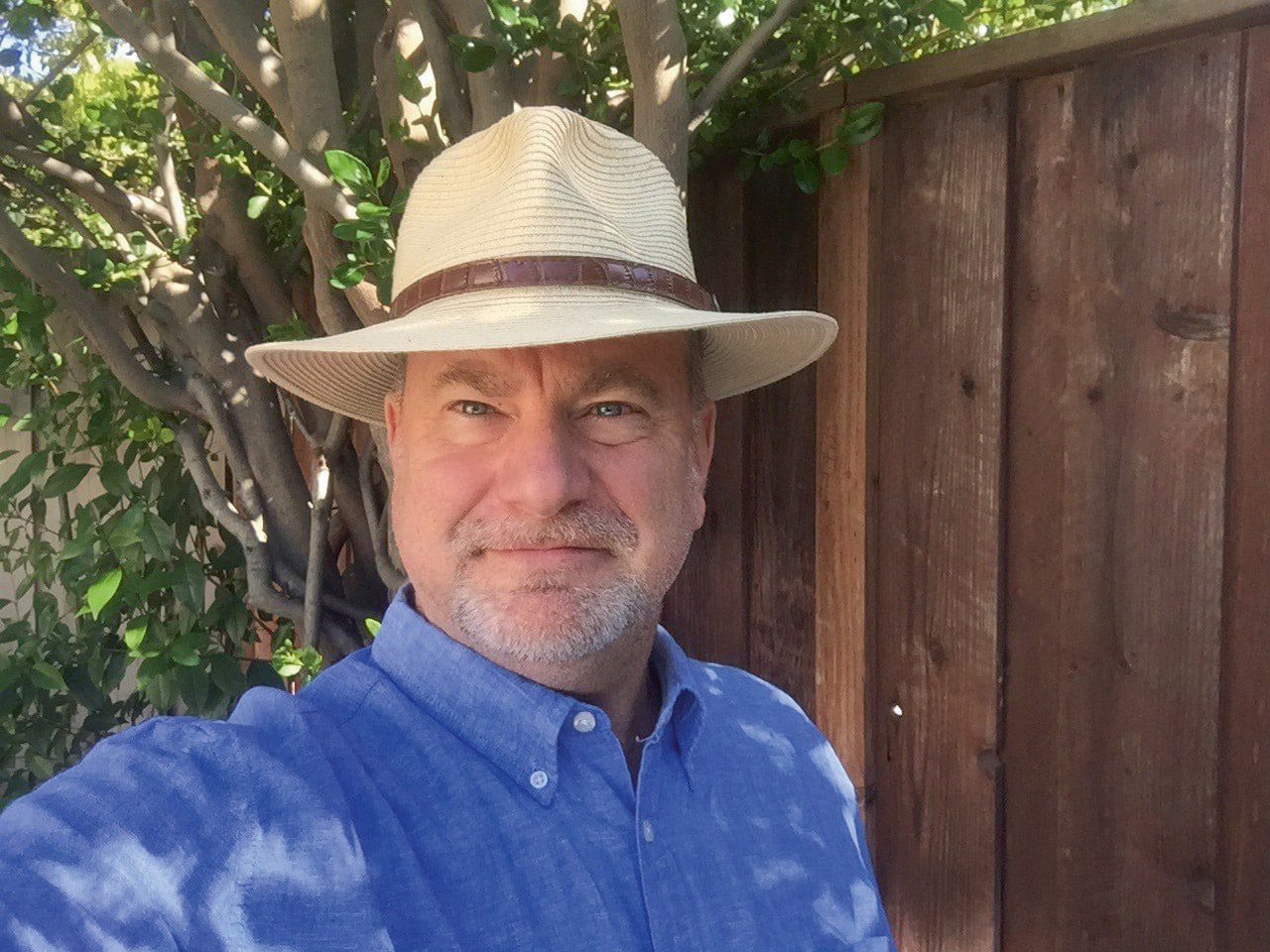a man wearing a lightcolored straw hat and a blue buttonup shirt is standing outdoors with green leaves and a wooden fence in the background digital production A man wearing a light-colored straw hat and a blue button-up shirt is standing outdoors, with green leaves and a wooden fence in the background.