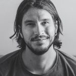 a smiling man with long hair and a beard wearing a gray tshirt seated in a casual setting with a neutral background digital production A smiling man with long hair and a beard, wearing a gray t-shirt, seated in a casual setting with a neutral background.