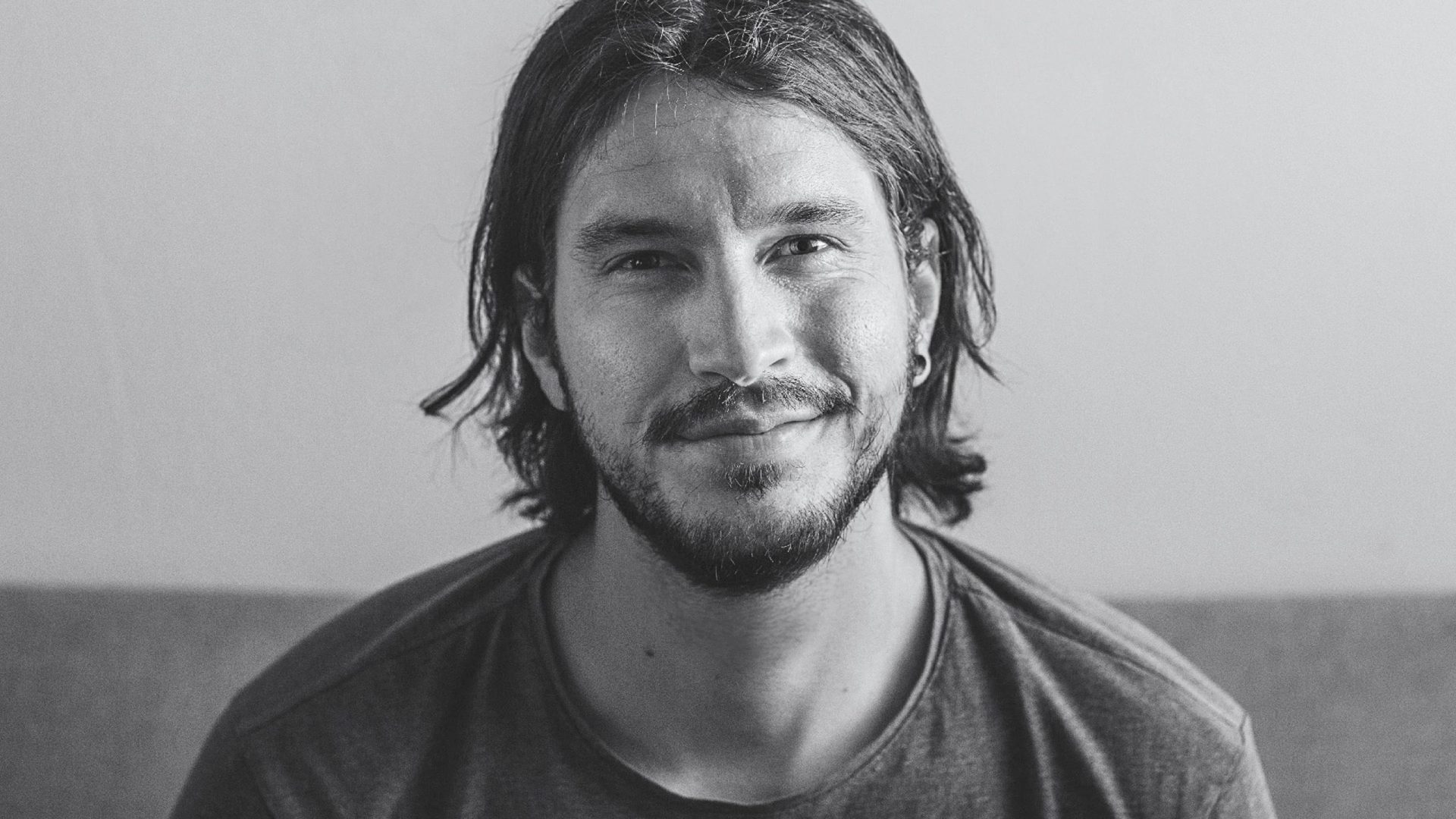 a smiling man with long hair and a beard wearing a gray tshirt seated in a casual setting with a neutral background digital production A smiling man with long hair and a beard, wearing a gray t-shirt, seated in a casual setting with a neutral background.