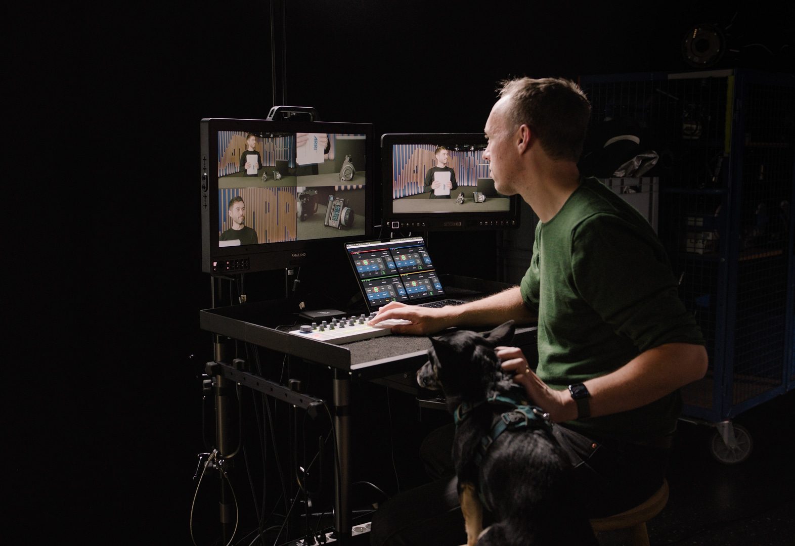 a man working at a desk with two monitors and a dog sitting beside him digital production A man working at a desk with two monitors and a dog sitting beside him.