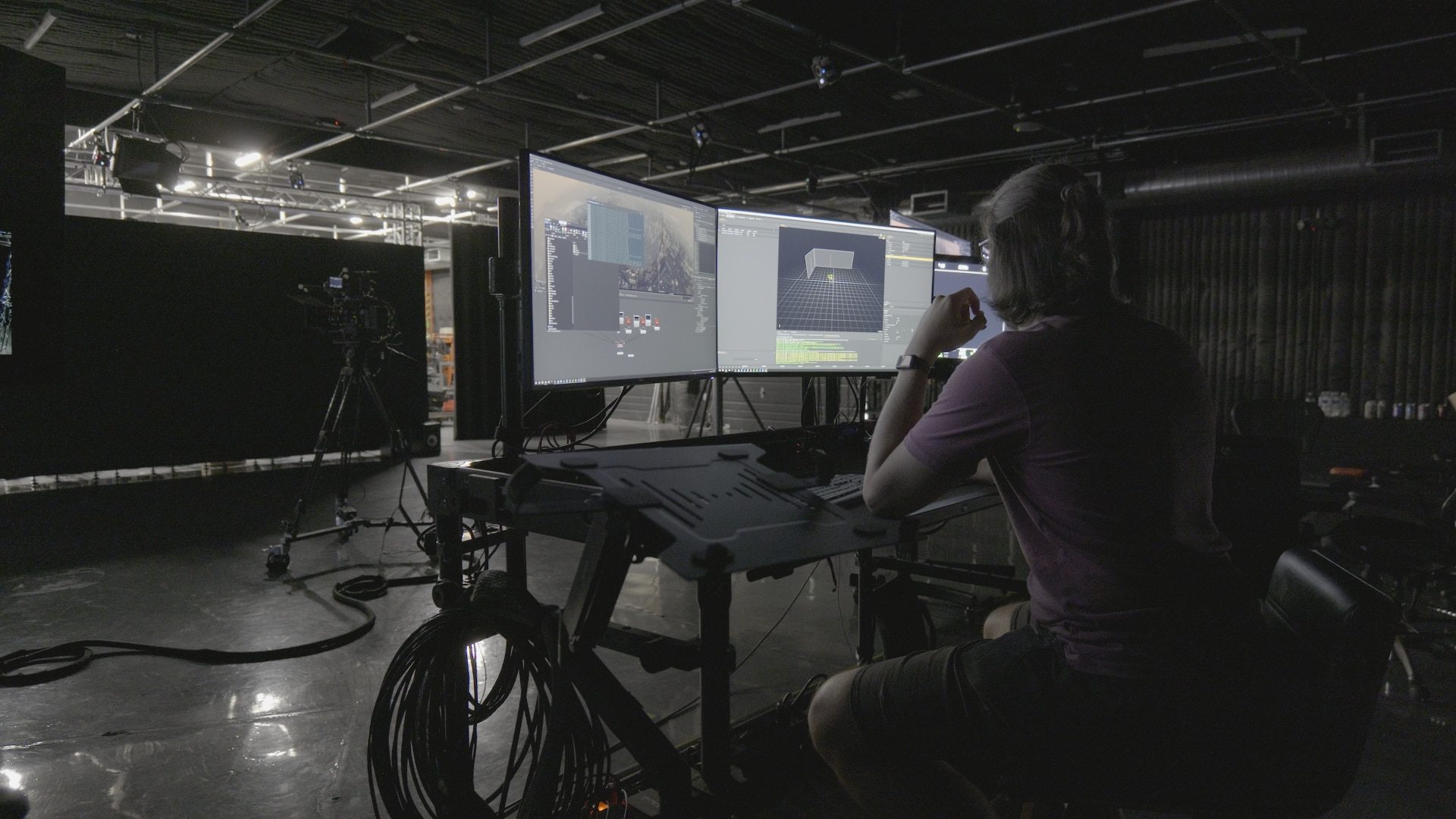 a person seated at a control desk with multiple computer screens displaying video editing software in a dimly lit studio cameras and equipment are visible in the background digital production A person seated at a control desk with multiple computer screens displaying video editing software in a dimly lit studio. Cameras and equipment are visible in the background.