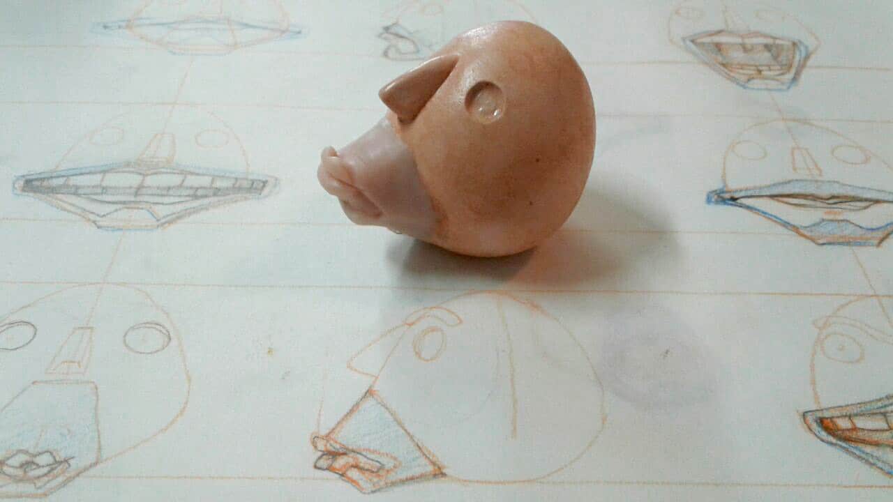 a clay model of a head with exaggerated features on a table covered with sketches of various face designs in light pencil and colored pencil the model is slightly offcenter with detail on one side digital production A clay model of a head with exaggerated features on a table covered with sketches of various face designs in light pencil and colored pencil. The model is slightly off-center with detail on one side.