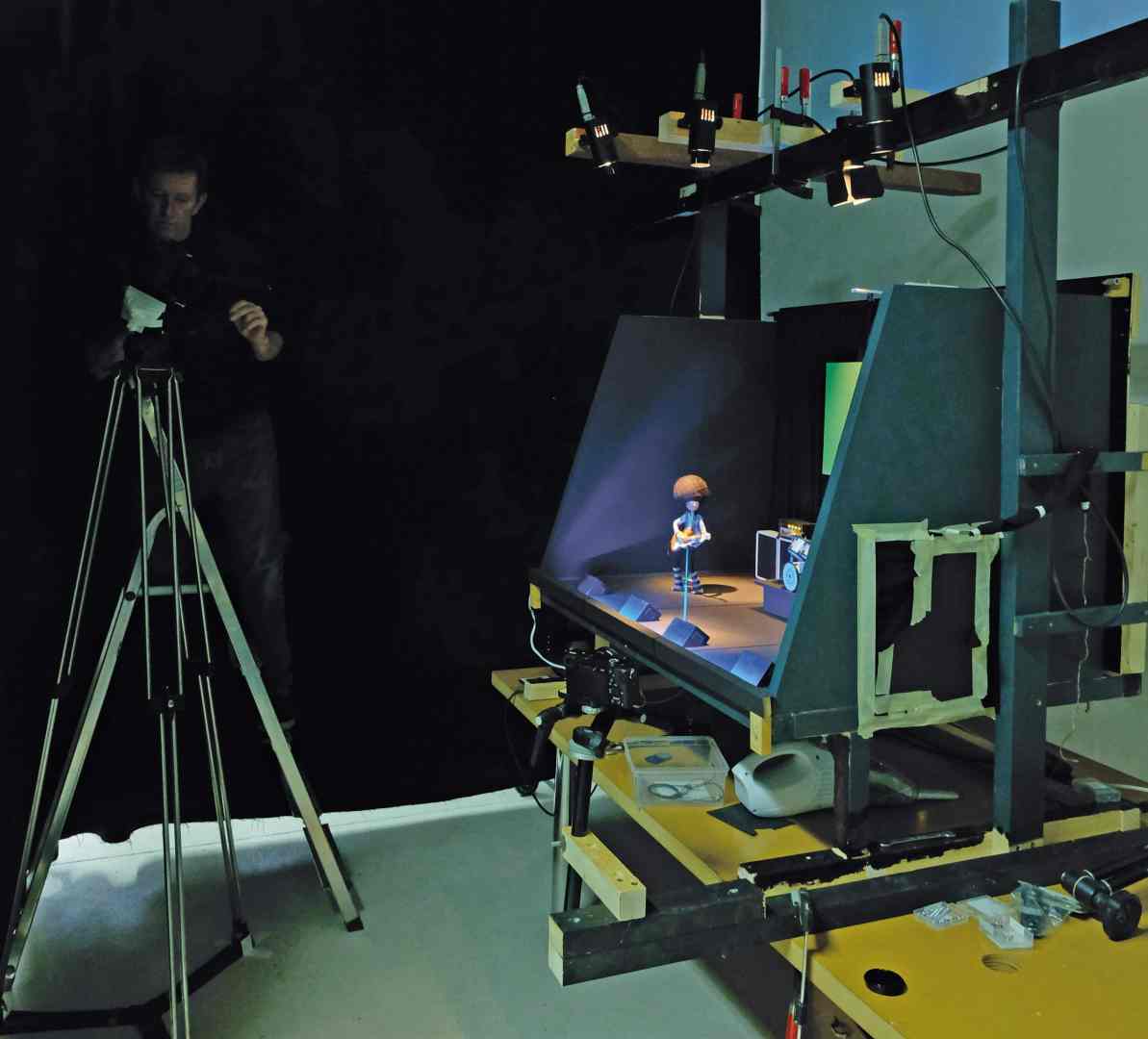 a person operating a camera on a tripod focusing on a small figurine displayed under studio lights the setup includes a backdrop and various equipment arranged on a wooden platform digital production A person operating a camera on a tripod, focusing on a small figurine displayed under studio lights. The setup includes a backdrop and various equipment arranged on a wooden platform.