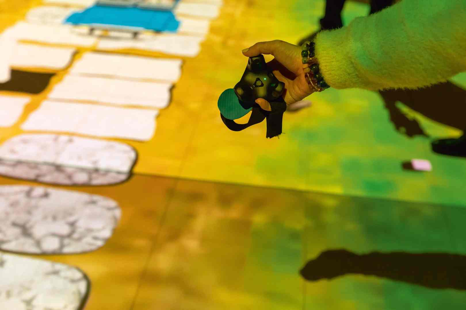 a hand holding a gaming controller over a colorful projected floor digital production A hand holding a gaming controller over a colorful projected floor.