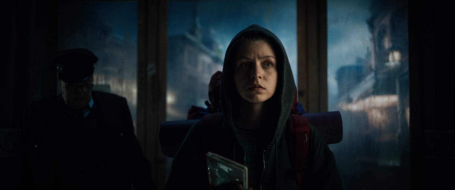 a young person wearing a hoodie stands in a dimly lit space looking intently forward while holding an item a person in uniform is visible in the background and the setting appears to be an outdoor area with a gloomy atmosphere digital production A young person wearing a hoodie stands in a dimly lit space, looking intently forward while holding an item. A person in uniform is visible in the background, and the setting appears to be an outdoor area with a gloomy atmosphere.
