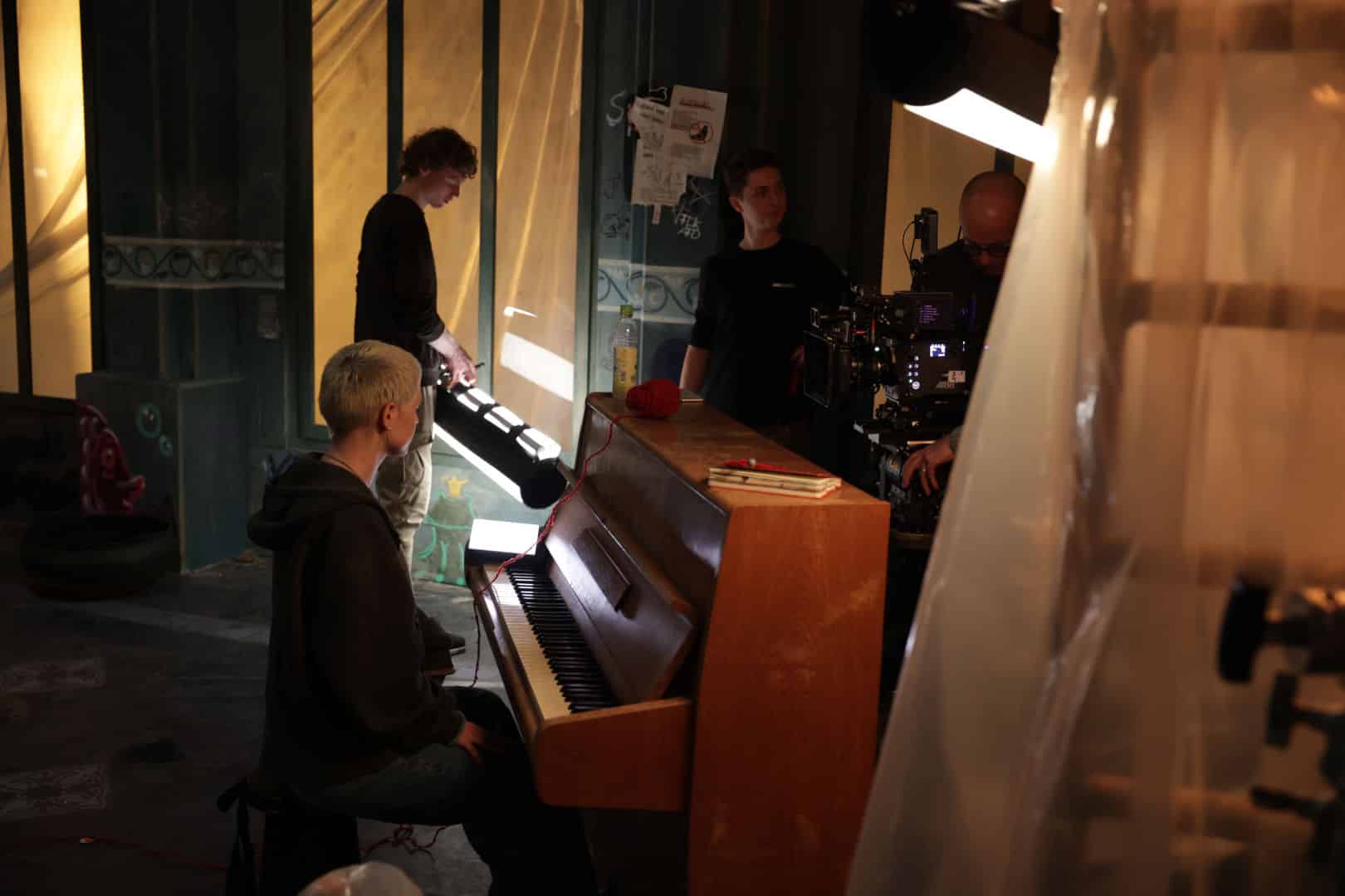 a musician playing a piano in a film set crew members are adjusting equipment and lighting in the background the set features green walls and natural light coming through windows digital production A musician playing a piano in a film set. Crew members are adjusting equipment and lighting in the background. The set features green walls and natural light coming through windows.