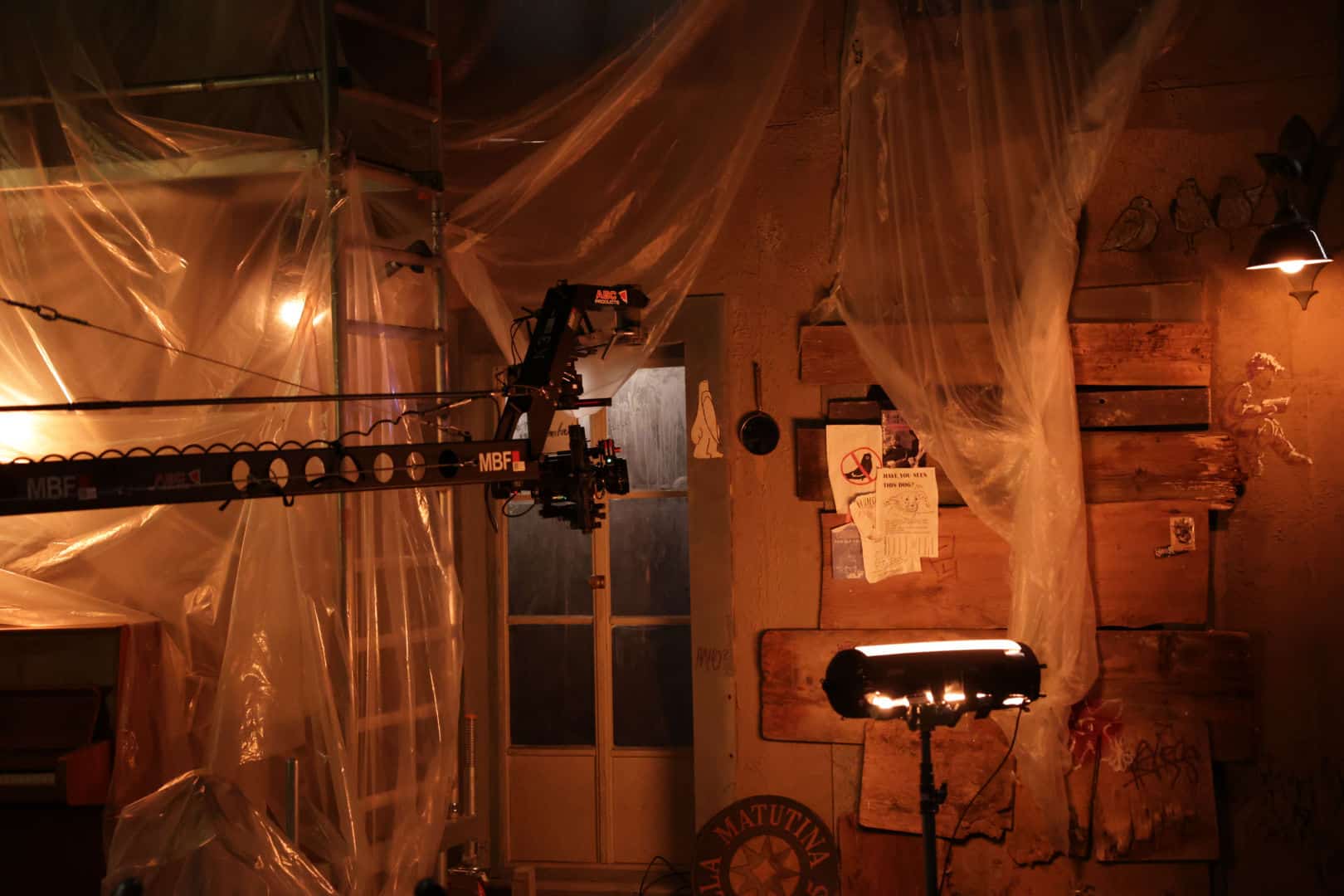 an interior space featuring a film set with plastic drapes hanging from the ceiling a camera crane and light stand are positioned in front of a wooden wall with various papers attached ambient lighting creates a moody atmosphere digital production An interior space featuring a film set with plastic drapes hanging from the ceiling. A camera crane and light stand are positioned in front of a wooden wall with various papers attached. Ambient lighting creates a moody atmosphere.