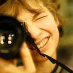 a person smiling while holding a camera close to their face with one eye looking through the viewfinder the background is blurred emphasizing the subject in focus digital production A person smiling while holding a camera close to their face, with one eye looking through the viewfinder. The background is blurred, emphasizing the subject in focus.