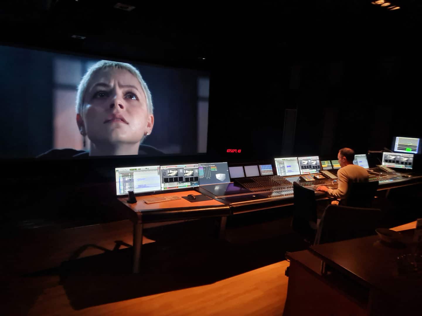 a person sitting in a dark editing room working at a control desk with multiple monitors while a large screen displays a closeup of a characters face with short blonde hair focused on a scene in a film digital production A person sitting in a dark editing room, working at a control desk with multiple monitors, while a large screen displays a close-up of a character's face with short blonde hair, focused on a scene in a film.