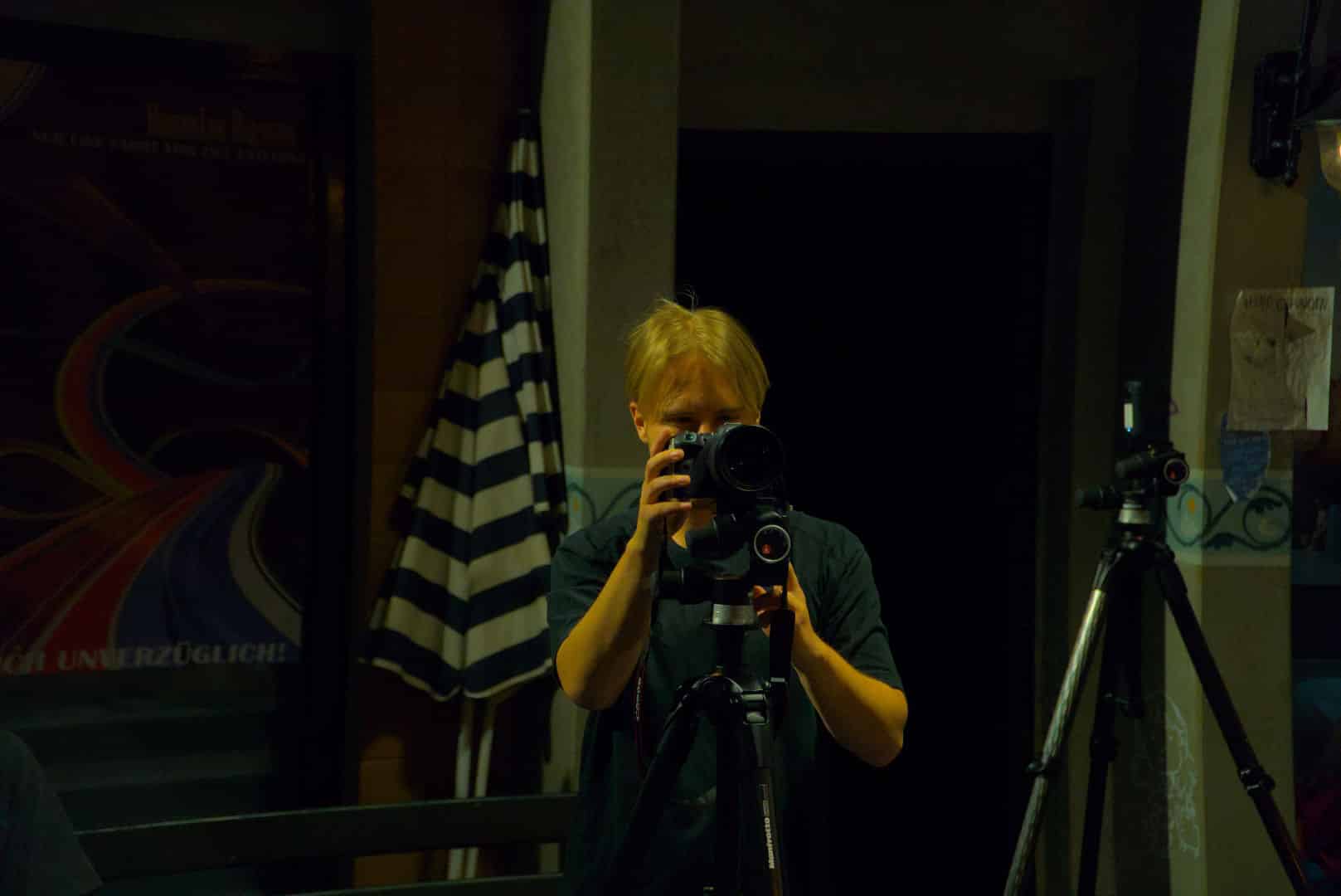 a person with blonde hair holding a camera looking through the viewfinder with a dark background and a striped flag hanging nearby two camera tripods stand next to them digital production A person with blonde hair holding a camera, looking through the viewfinder, with a dark background and a striped flag hanging nearby. Two camera tripods stand next to them.
