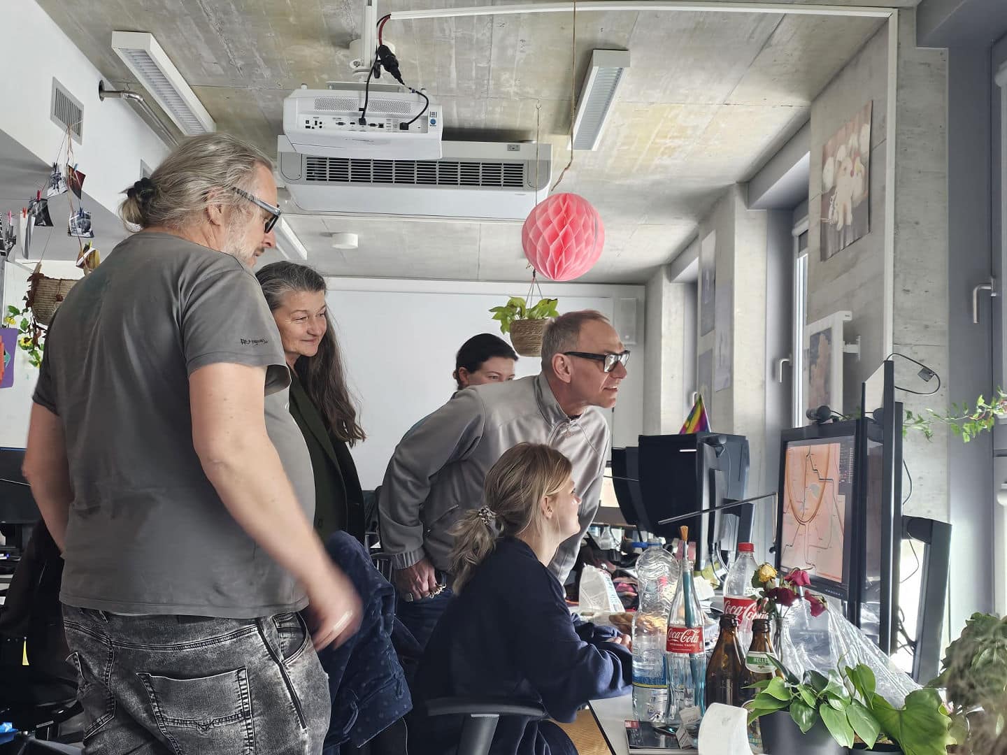 a group of four people observing a computer screen in an office environment with plants and personal items on desks soft lighting enhances the workspaces atmosphere digital production A group of four people observing a computer screen in an office environment, with plants and personal items on desks. Soft lighting enhances the workspace's atmosphere.