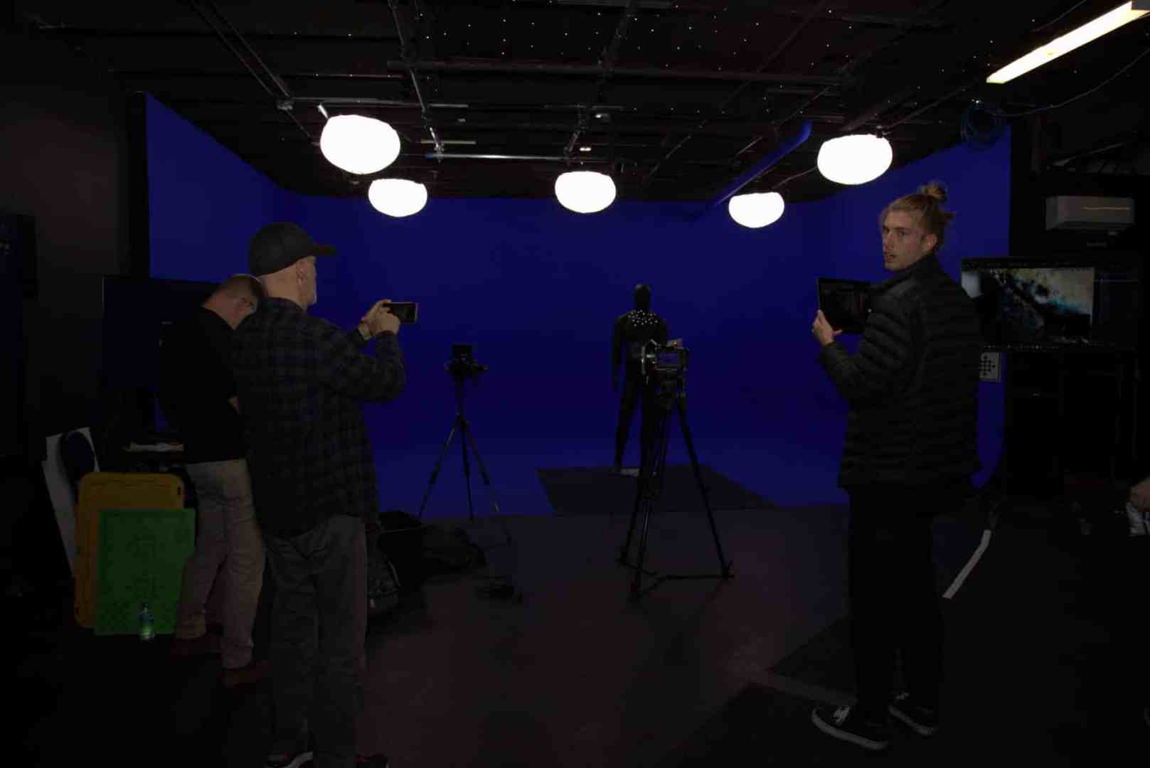 two men in a studio with a blue backdrop one holding a camera and adjusting settings while the other poses with a light meter next to a mannequin studio lights hang from the ceiling illuminating the scene digital production Two men in a studio with a blue backdrop, one holding a camera and adjusting settings, while the other poses with a light meter next to a mannequin. Studio lights hang from the ceiling, illuminating the scene.
