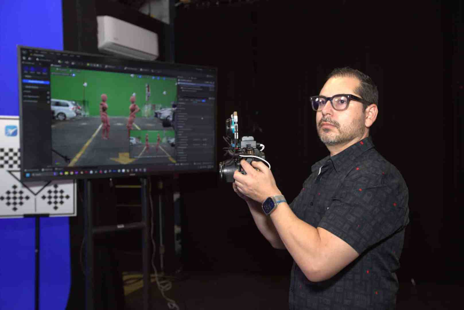a person holding a camera while standing in front of a monitor displaying a green screen project featuring animated figures the background is dark and there are technical elements visible on the screen digital production A person holding a camera while standing in front of a monitor displaying a green screen project featuring animated figures. The background is dark, and there are technical elements visible on the screen.