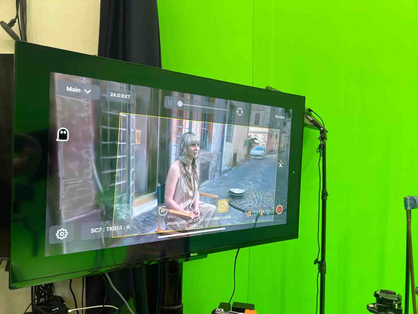 a display screen showing a live capture of a woman sitting in a street setting with a green backdrop visible behind the screen the screen shows controls and settings for video capture digital production A display screen showing a live capture of a woman sitting in a street setting, with a green backdrop visible behind the screen. The screen shows controls and settings for video capture.