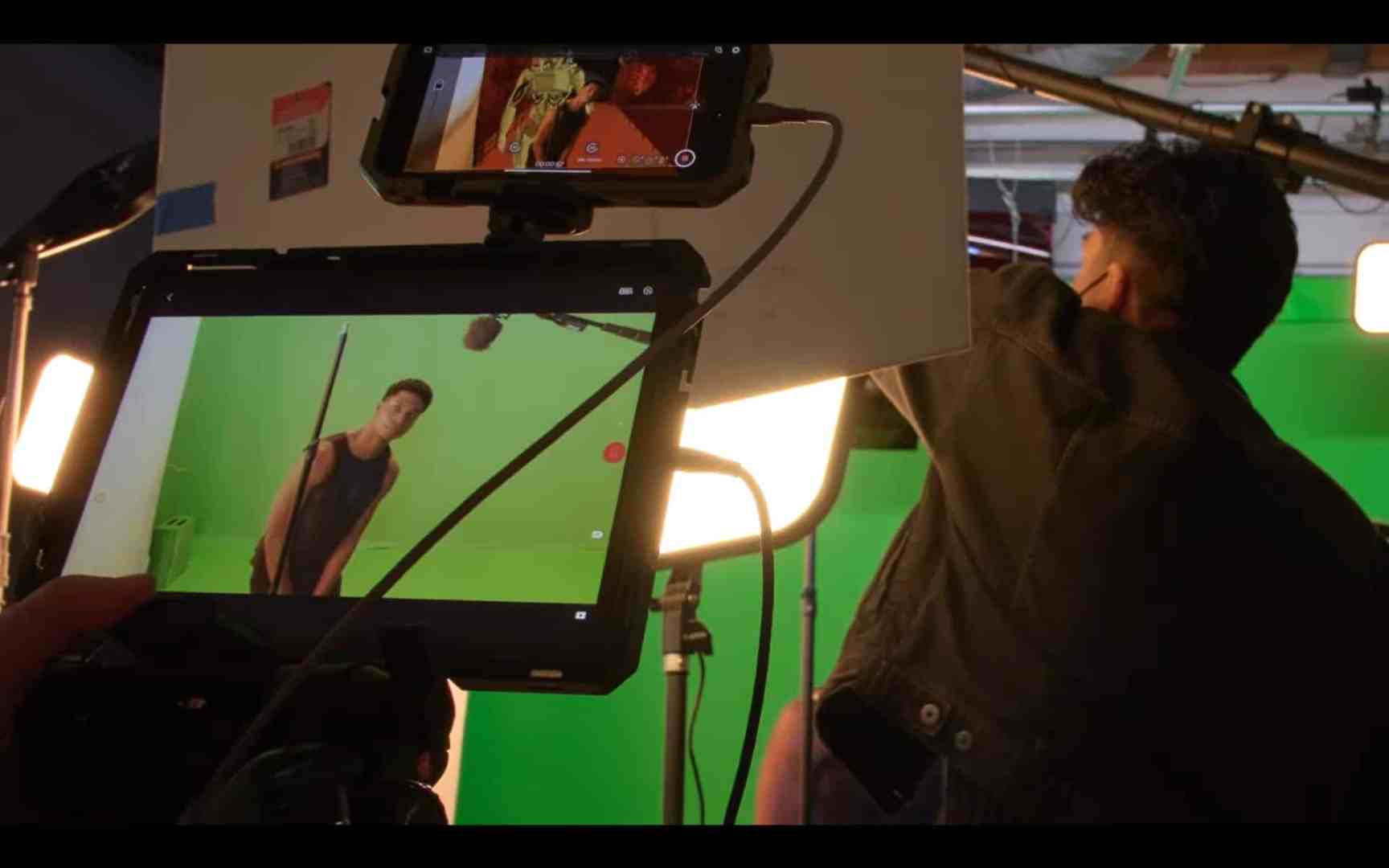 a tablet screen shows a performer in a studio with a green backdrop while a crew member adjusts lighting equipment in the background the setting is illuminated with various studio lights digital production A tablet screen shows a performer in a studio with a green backdrop, while a crew member adjusts lighting equipment in the background. The setting is illuminated with various studio lights.