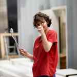 a person wearing a red tshirt and blue jeans stands in a workspace holding a stick with a small item on the end and giving a thumbsup gesture the background features blurred furniture and equipment digital production A person wearing a red t-shirt and blue jeans stands in a workspace, holding a stick with a small item on the end and giving a thumbs-up gesture. The background features blurred furniture and equipment.