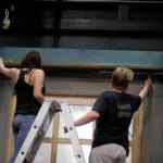 two individuals working on a stage set one person is standing on a ladder painting a wall while the other is reaching up to touch the structure they are both dressed casually with one wearing a black tank top and the other a black tshirt digital production Two individuals working on a stage set. One person is standing on a ladder painting a wall, while the other is reaching up to touch the structure. They are both dressed casually, with one wearing a black tank top and the other a black T-shirt.