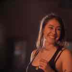 a woman smiling while holding a small glass wearing a black tank top with soft lighting creating a warm ambiance the setting appears casual indicating a relaxed atmosphere digital production A woman smiling while holding a small glass, wearing a black tank top, with soft lighting creating a warm ambiance. The setting appears casual, indicating a relaxed atmosphere.