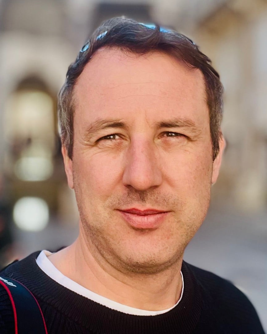 a man with short dark hair smiling at the camera in a blurred outdoor background digital production A man with short dark hair smiling at the camera in a blurred outdoor background.