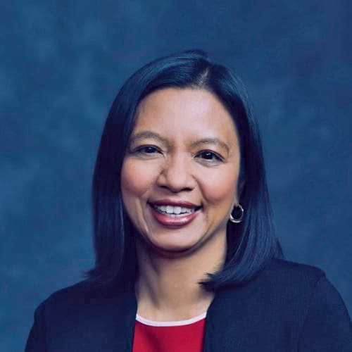 a woman smiling with straight black hair wearing a black blazer over a red top against a dark background digital production Headshot of a woman with shoulder-length dark hair, wearing a black blazer over a red top, smiling against a blue background.