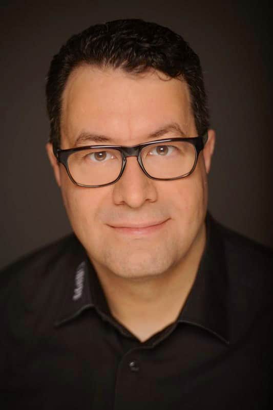 man with glasses smiling in a dark shirt digital production Portrait of a professional man wearing glasses and a black shirt with a slight smile on his face against a dark background.