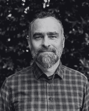 a man with a beard wearing a checkered shirt standing outdoors with foliage in the background digital production A man with a beard wearing a checkered shirt, standing outdoors with foliage in the background.