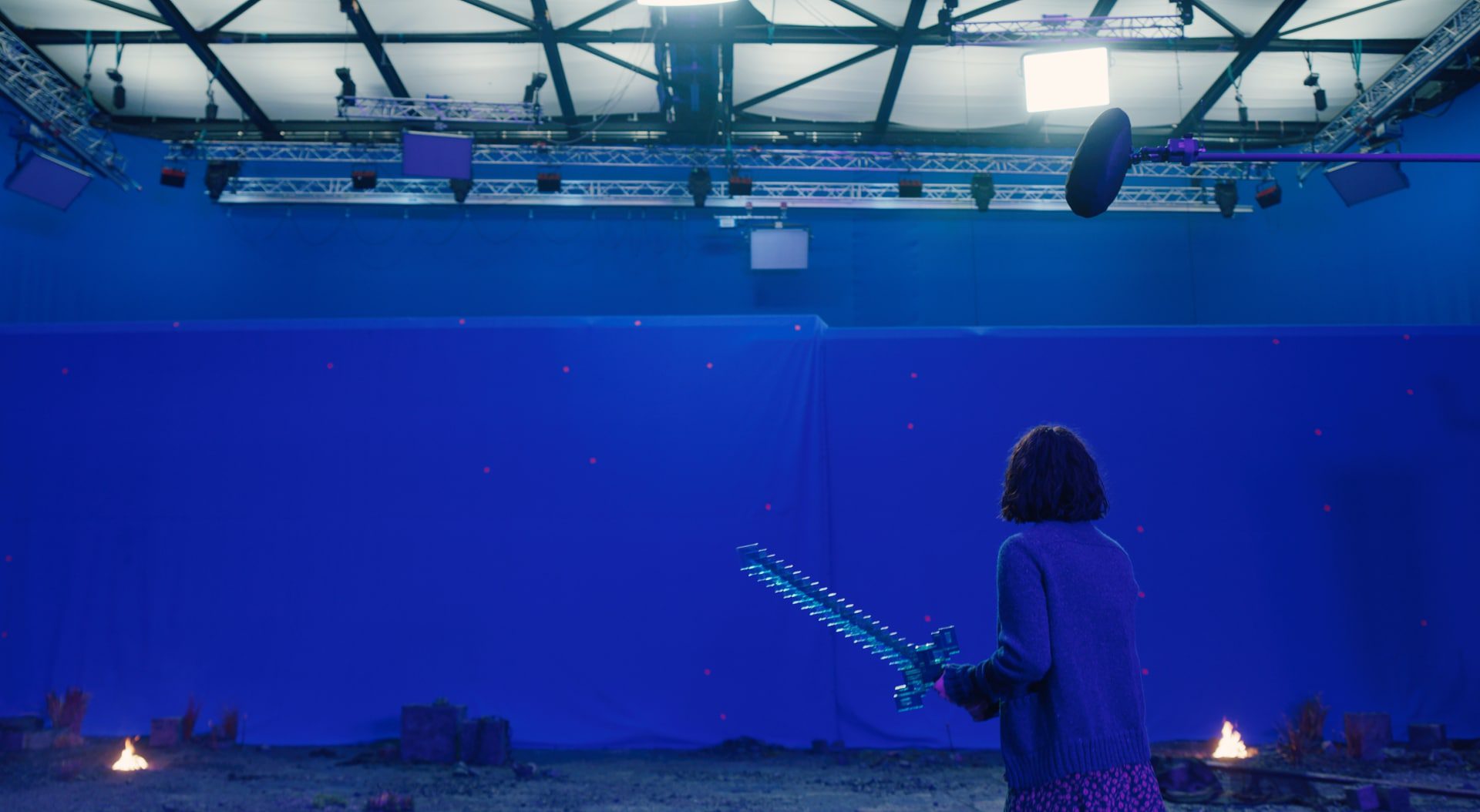 a person holding a sword in a filming studio with blue backdrop digital production A person holding a prop sword stands in a blue screen studio, with scattered debris and a small fire in the foreground.