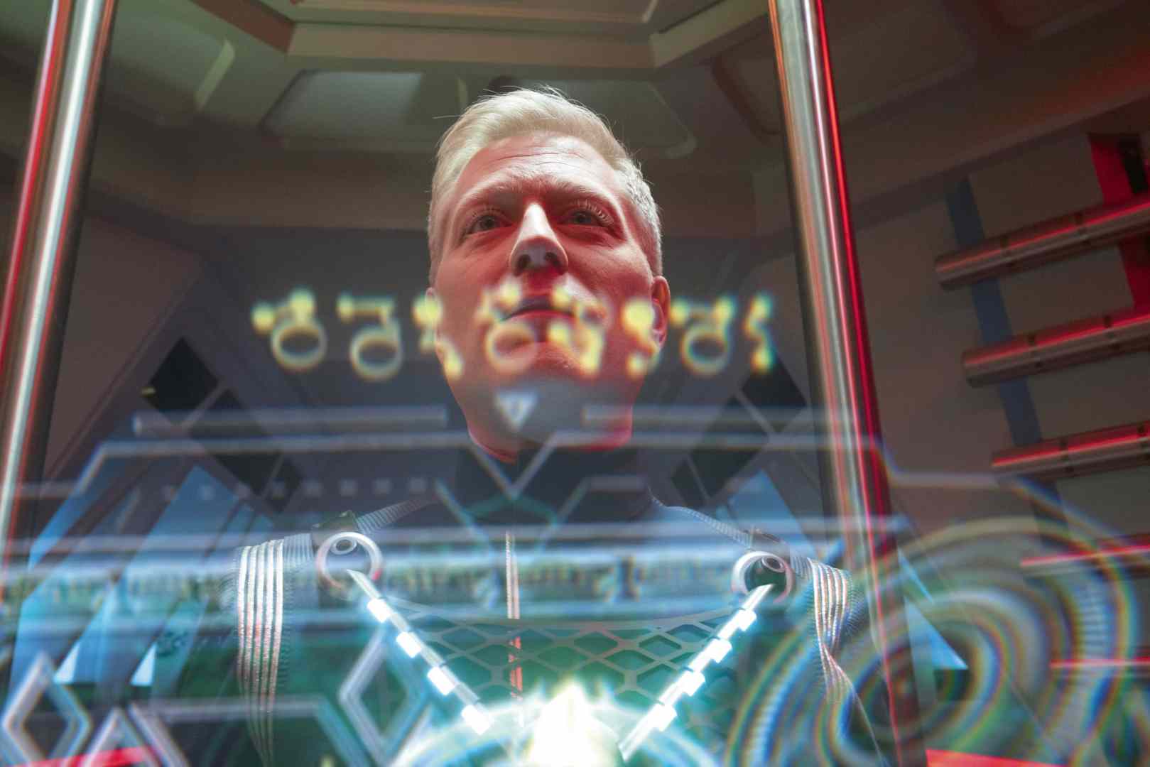 a closeup of a man in a spacethemed suit standing inside a futuristic glass chamber holographic displays with digital symbols are visible in the foreground casting an ambient glow the background features sleek metallic designs and red lighting digital production A close-up of a man in a space-themed suit, standing inside a futuristic glass chamber. Holographic displays with digital symbols are visible in the foreground, casting an ambient glow. The background features sleek metallic designs and red lighting.