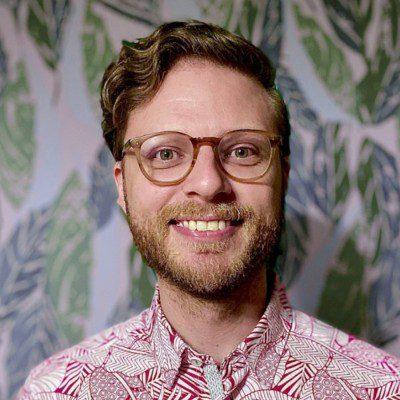 a man with curly hair and glasses smiles at the camera while wearing a colorful patterned shirt the background features a leafy design with various shades of green digital production A man with curly hair and glasses smiles at the camera while wearing a colorful patterned shirt. The background features a leafy design with various shades of green.