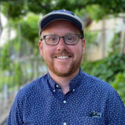 a smiling man with glasses and a cap wearing a blue patterned shirt stands outdoors in a green leafy setting he appears friendly and approachable with lush greenery in the background digital production A smiling man with glasses and a cap, wearing a blue patterned shirt, stands outdoors in a green, leafy setting. He appears friendly and approachable, with lush greenery in the background.